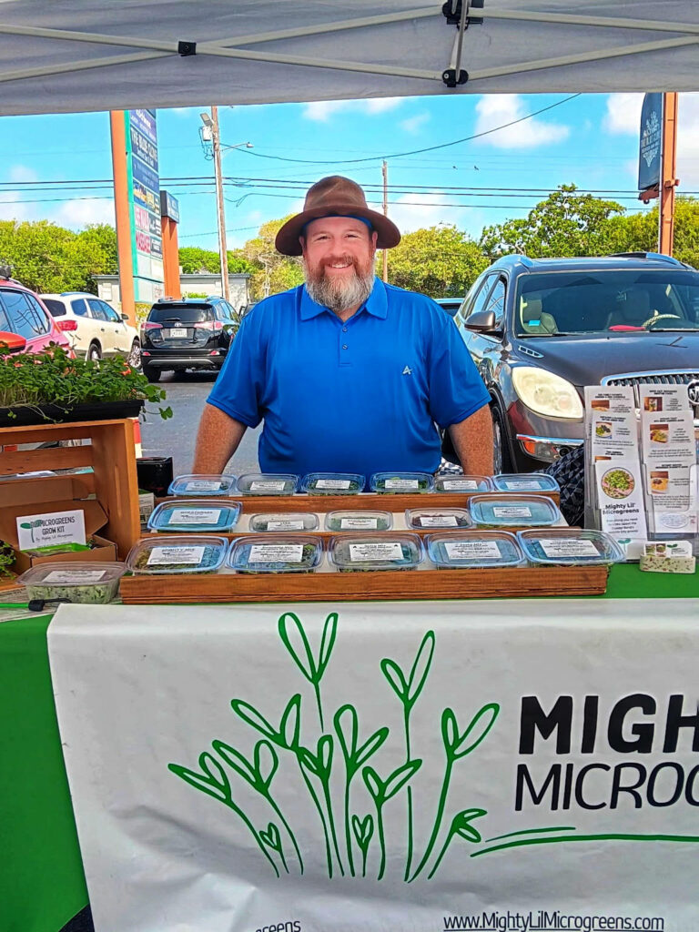 A picture of a man in a blue shirt standing at a market table with microgreens in front of him.